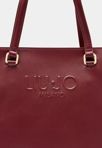 Raudona tekstūruota rankinė su įspauduotu "LIU•JO MILANO" logotipu, turinti dvi rankenas ir auksinės spalvos detalių akcentus.