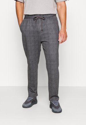 Mężczyzna w szarych spodniach w kratę ze ściągaczem i bocznymi kieszeniami oraz w ciemnoszarych butach sportowych, stoi na tle jednolitej ściany.