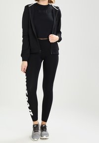 Person wearing black Nike hoodie, cropped logo t-shirt, black leggings with white text, and gray Nike sneakers, standing against a neutral background.