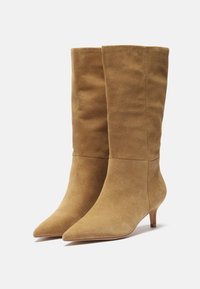 Bottes en suedine beige à bout pointu et hauteur jusqu'au genou, avec une texture lisse et un talon slim et bas. Elles présentent un tige ajustée et un design épuré et minimal.