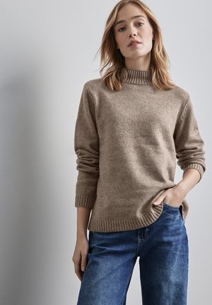 Beige knitted sweater with a ribbed high neckline and cuffed sleeves, worn over blue denim jeans. Textured fabric with a relaxed fit.