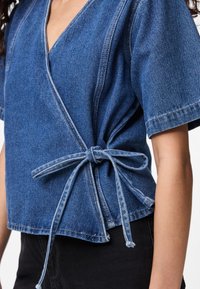 Top avvolgente in denim con scollatura a V profonda, maniche corte, dettagli cuciti e chiusura con nodo laterale. Colore: blu medio. Finitura testurizzata.