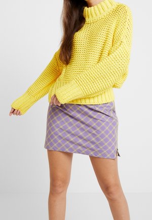 Femme portant un gros pull en maille jaune associé à une mini-jupe à carreaux violette taille haute avec une fermeture éclair latérale.