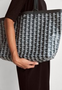 Personne tenant un sac fourre-tout noir avec un motif géométrique blanc et des poignées doubles, vêtue de vêtements foncés sur un fond uni.