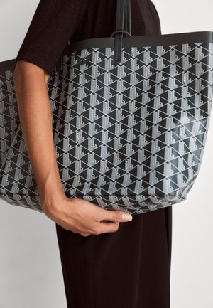 Personne tenant un sac fourre-tout noir avec un motif géométrique blanc et des poignées doubles, vêtue de vêtements foncés sur un fond uni.