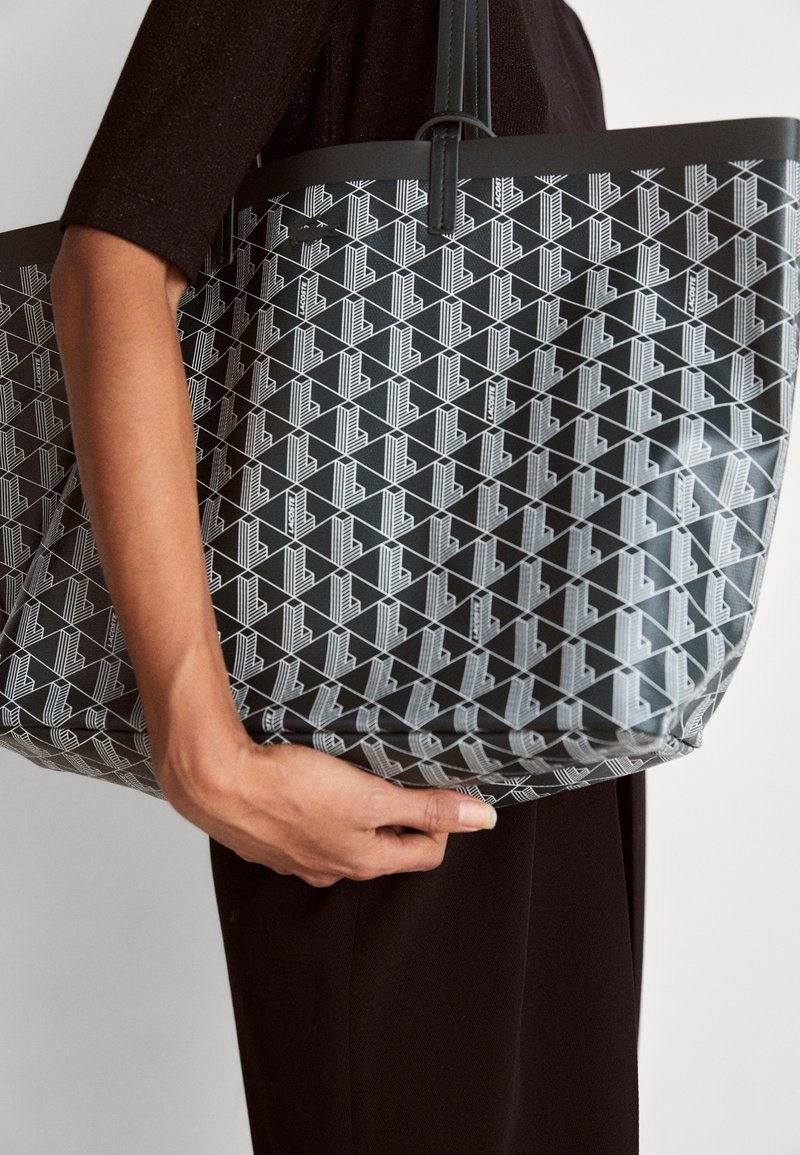 Personne tenant un sac fourre-tout noir avec un motif géométrique blanc et des poignées doubles, vêtue de vêtements foncés sur un fond uni.