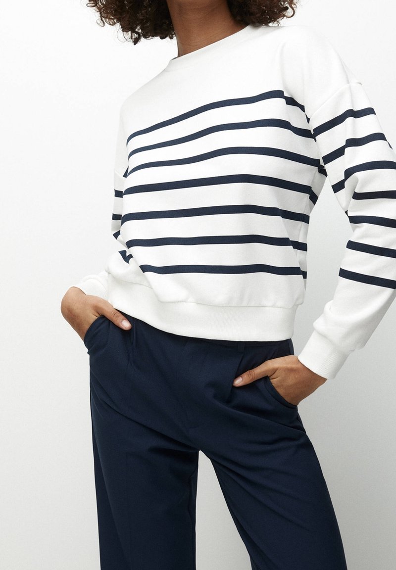 Personne portant une chemise blanche à manches longues avec des rayures horizontales bleu marine et un pantalon bleu marine foncé, les mains dans les poches, sur un fond uni.