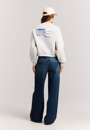 Person wearing a light gray sweatshirt with blue text on the back, blue wide-leg jeans, and a beige baseball cap, standing facing away.