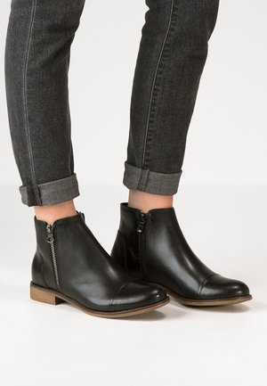 Ankelhøje sorte læderstøvler med lynlåse i siden, båret med mørkegrå optrukne jeans mod en ensfarvet hvid baggrund.