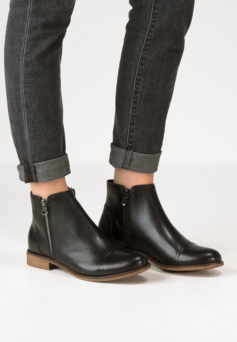 Bottes en cuir noires montant jusqu'à la cheville avec des fermetures éclair sur le côté, portées avec un jean gris foncé retroussé, sur un fond blanc uni.
