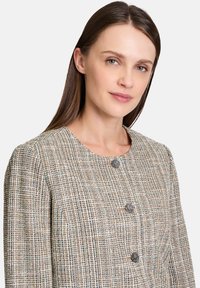 Tweed jacket in beige with a textured grid pattern. Features round neckline and three dark buttons at the front.