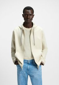  lys beige zip-up hoodie med hætte og snore, en cremet sweater under og blå denimjeans. Har ensfarvede nuancer og subtil tekstur.