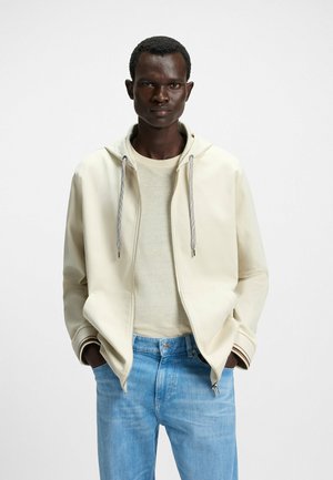Ljusbeige zip-up hoodie med dragsko på huvan, krämfärgad tröja under och blå denimjeans. Har enhetliga färger och subtil textur.
