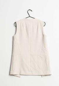 Gilet sans manches beige sur un cintre noir. Présente une coupe nette, une couture au dos et deux fentes latérales à l'ourlet. Texture du tissu douce.