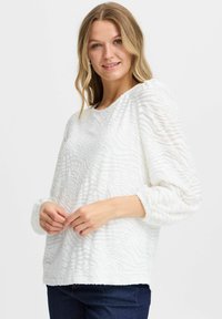 Vit texturerad långärmad topp med ett vågigt mönster, rund halsringning och elastiska manschetter. Matchad med mörkblå jeans.