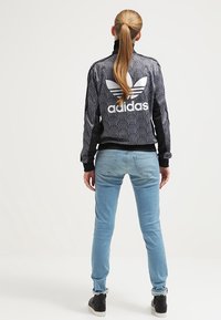 Černá zipová bunda s bílým logem Adidas a vroubkovaným vzorem na zadní straně, doplněná o světle modré džíny a černé tenisky.