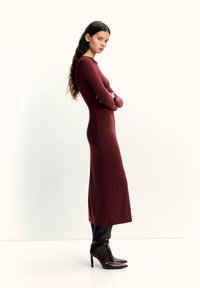 Rochie visinie cu mâneci lungi, din material elastic, siluetă mulată, cu tiv până la genunchi, asortată cu cizme negre lucioase cu toc stiletto până la genunchi.