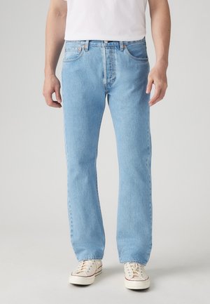 Homme portant un jean droit bleu clair, des baskets blanches et un t-shirt blanc, debout devant un fond clair uni.