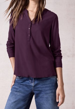 Femme portant un chemisier violet foncé à manches longues avec trois boutons et un jean en denim bleu, debout devant un fond clair.
