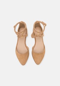 Un par de bailarinas de gamuza color beige con punta afilada, con dobles correas en el tobillo y pequeñas hebillas doradas, vistas desde arriba sobre un fondo blanco.