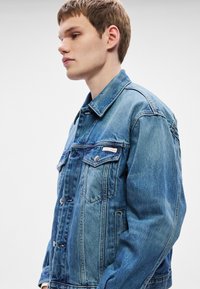 Μπουφάν από τζιν σε ανοιχτό μπλε χρώμα, με κουμπιά κλεισίματος, δύο τσέπες στο στήθος και ετικέτα στην αριστερή τσέπη. Ορατές λεπτομέρειες ραφής.