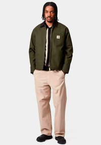 Uomo con capelli intrecciati indossa una giacca verde oliva con zip, una maglietta nera, pantaloni beige e scarpe nere, in piedi con le mani nelle tasche.