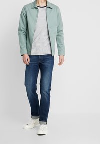 Muntgroene lichtgewicht jas over een grijs gestreept T-shirt, gecombineerd met donkerblauwe jeans en witte sneakers. Eenvoudig ontwerp en casual pasvorm.