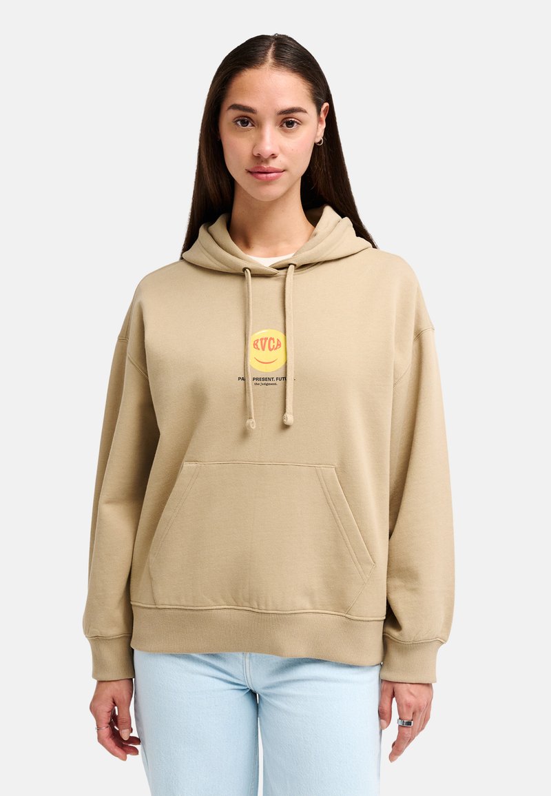 Mujer con cabello largo y oscuro, lleva una sudadera beige con un logo amarillo de carita sonriente y jeans azul claro, de pie contra un fondo blanco.