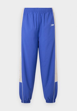 Pantaloni sportivi blu con pannelli laterali beige, fascia elastica in vita e polsini alla caviglia, con un piccolo logo bianco sulla parte superiore della coscia.