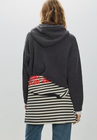 Sötétszürke vastag kötött kapucnis pulóver, alatta fekete-fehér csíkos ing piros felirattal a derékrésznél. Laza szabás.