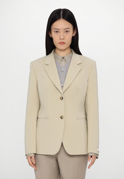 Beige tailored blazer with two front buttons, notch lapels, and side pockets; worn over a grey button-up shirt. Smooth fabric, structured shape.