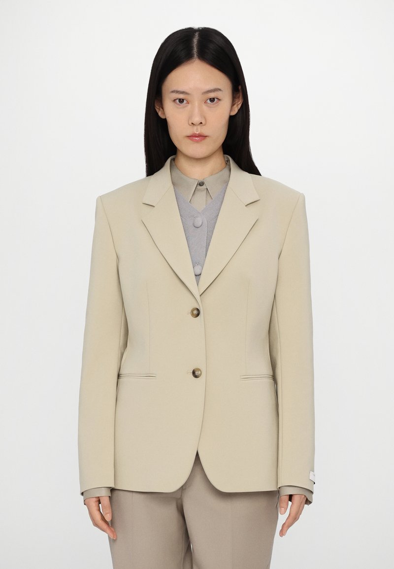 Beige tailored blazer with two front buttons, notch lapels, and side pockets; worn over a grey button-up shirt. Smooth fabric, structured shape.
