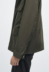 Olivengrøn, letvægtsjakke med en glat tekstur, sidelommer og en knapdetalje ved manchet, vist fra siden.