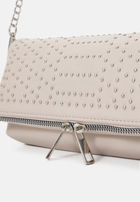 Pochette en cuir beige avec des clous argentés formant un motif géométrique, fermeture éclair double argentée et bandoulière en chaîne argentée.