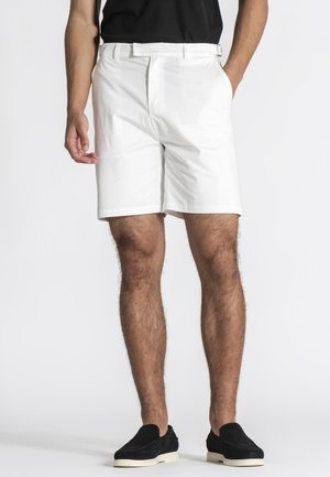 Muškarac u bijelim kratkim hlačama do koljena, crnim slip-on cipelama i crnoj majici, stoji s rukama u džepovima na neutralnoj pozadini.