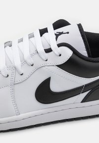 Białe skórzane sneakersy z czarnymi akcentami, charakteryzujące się wyraźnym czarnym logo, fakturowanym językiem oraz białymi sznurowadłami. Gumowa podeszwa z wzorem zapewniającym przyczepność.