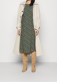 Beige trenchcoat, grön blommönstrad klänning och tan knähöga stövlar. Har knappar, långa ärmar och en flödande design. Släta texturer genomgående.