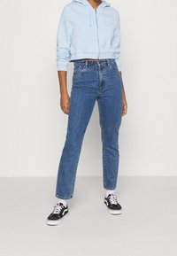 Ljusblå cropped hoodie med dragkedja, kombinerad med högmidjade blå jeans. Modellen bär svarta och vita sneakers. Enkel, avslappnad stil.