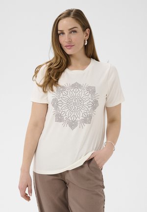 Kobieta o długich brązowych włosach, ubrana w białą bluzkę z krótkim rękawem zdobioną kropkowanym kwiatowym wzorem oraz taupe spodnie, pozująca z jedną ręką w kieszeni.