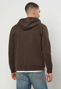Homme portant un sweat à capuche marron foncé et un jean bleu clair, debout de dos devant un fond clair uni.