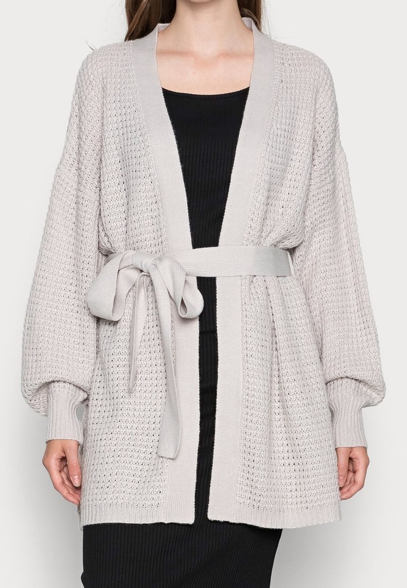Femme portant un cardigan ajouré gris clair avec de larges poignets côtelés et une ceinture nouée sur une robe noire côtelée.