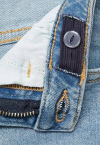 Tessuto denim di colore azzurro chiaro con cuciture gialle a contrasto, una placchetta dei bottoni a coste blu navy e una zip metallica. Dettaglio ravvicinato delle cuciture.
