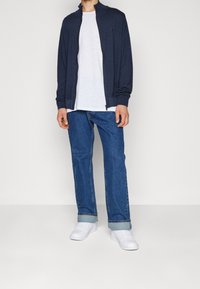 Man som bär en mörkblå zip-up-jacka, vit t-shirt, uppvikta blå jeans och vita sneakers som står mot en enkel vit bakgrund.