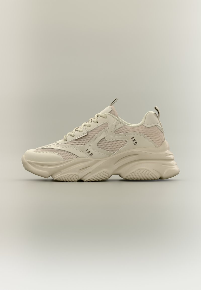 Zapatilla beige de estilo grueso con suela gruesa, diseño con cordones, paneles de malla y cuero, y tiradores en la lengüeta y el talón, mostrada de perfil.