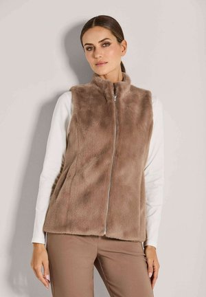 Vrouw in een lichtbruine nepbont zip-up vest over een witte langslavend top en bijpassende lichtbruine broek, staande tegen een effen muur.