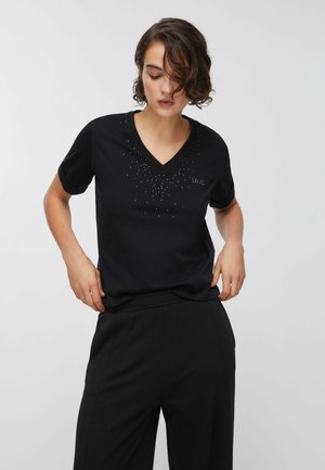 Donna con una T-shirt nera a V con piccole perline decorative, abbinata a pantaloni neri a gamba larga, su uno sfondo chiaro e semplice.