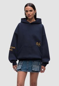 Felpa navy con ampio tascone frontale, dettagli ricamati in giallo e vestibilità over, abbinata a una gonna di jeans con tasche.