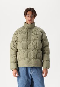 Olivengrønn pufferjakke med høy krage, kviltet design, frontlås og subtil merking. Kombinert med lyseblå denimbukser.
