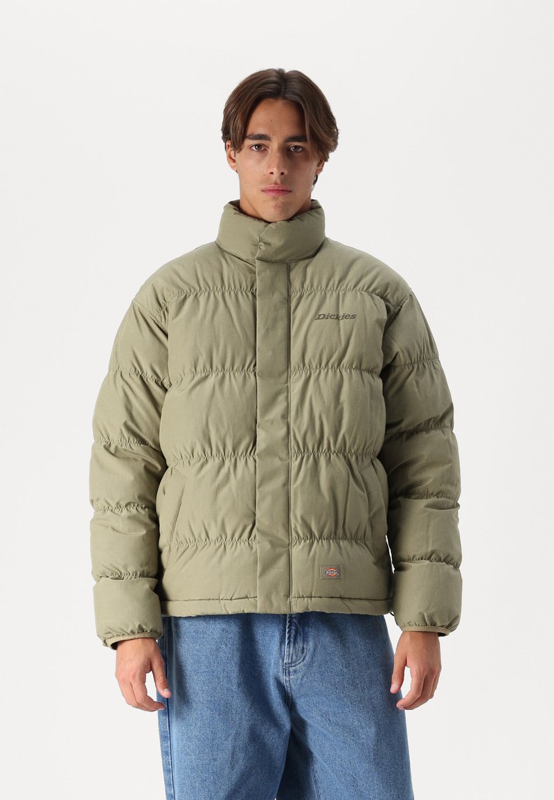 Olivengrønn pufferjakke med høy krage, kviltet design, frontlås og subtil merking. Kombinert med lyseblå denimbukser.