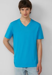 T-shirt a manica corta con scollo a V di colore blu brillante, realizzato in tessuto morbido. Presenta un design semplice con spacchi laterali all'orlo per facilitare i movimenti.